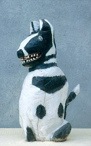 A small, roughly carved dog sculpture painted white with black patches, sitting upright with visible teeth and a slightly worn, handmade appearance against a plain background.