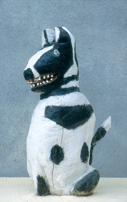 A small, roughly carved dog sculpture painted white with black patches, sitting upright with visible teeth and a slightly worn, handmade appearance against a plain background.