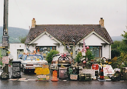 A small house with numerous handmade signs, objects, and decorations cluttering the front yard, driveway, and cars parked outside.