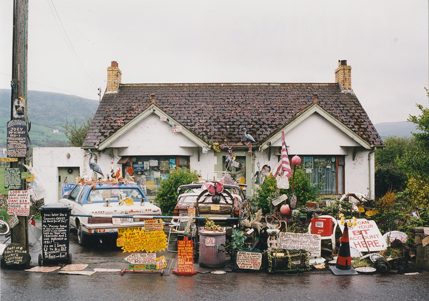 A small house with numerous handmade signs, objects, and decorations cluttering the front yard, driveway, and cars parked outside.
