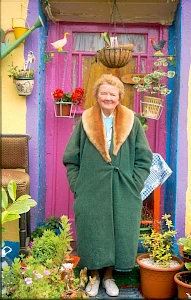 Elderly woman stands smiling in front of a colorful, flower-adorned doorway. She wears a green coat with a fur collar, exuding warmth and cheerfulness.