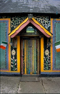 A colorful mosaic wall surrounds a vividly painted door with vertical stripes. Irish flags flank the entrance, creating a vibrant and artistic scene.