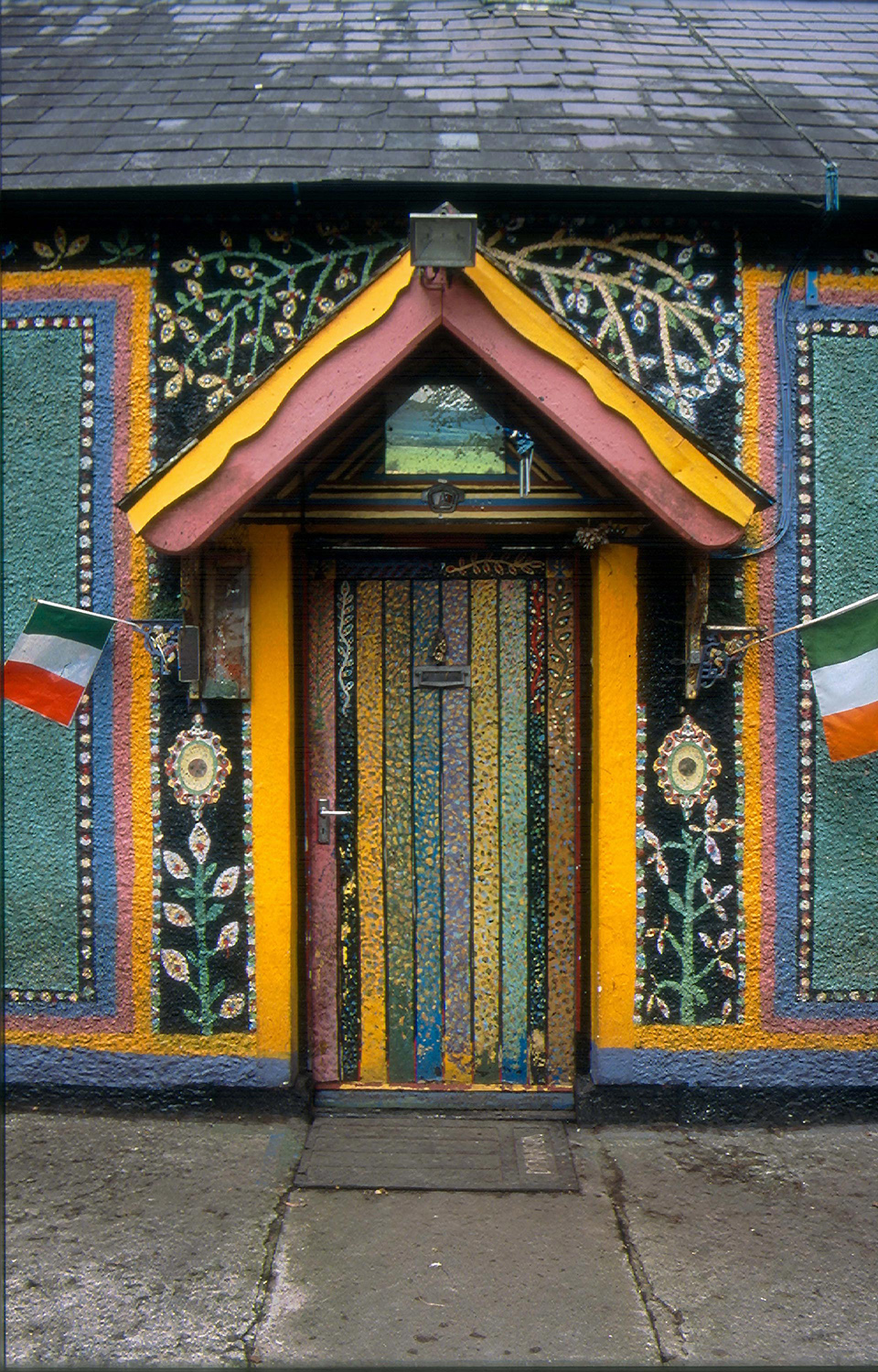 A colorful mosaic wall surrounds a vividly painted door with vertical stripes. Irish flags flank the entrance, creating a vibrant and artistic scene.